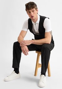 Schwarz gestreifter Jumpsuit über einem weißen Poloshirt, kombiniert mit weißen Sneakers. Das Modell sitzt auf einem Holzschemel und präsentiert das geschichtete Design des Outfits.