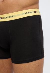 Κοντινό πλάνο ατόμου που φοράει μαύρα ανδρικά εσώρουχα τύπου boxer με κίτρινη μέση, διακοσμημένη με την επωνυμία «Tommy Hilfiger» και το λογότυπο.