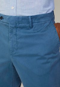 Jasnoniebieskie bawełniane spodnie o gładkiej strukturze, z płaskim przodem, szlufkami na pasek oraz brązowym akcentem na tylnej kieszeni.