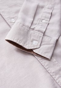 Ljust beige knapp-up skjorta i texturerat tyg, med krage och små vita knappar. Tyget har ett diskret mönster.