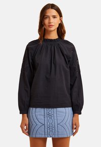 Blusa nera in cotone con scollatura arricciata e dettagli floreali ricamati sulle maniche, abbinata a una gonna trapuntata blu chiaro.