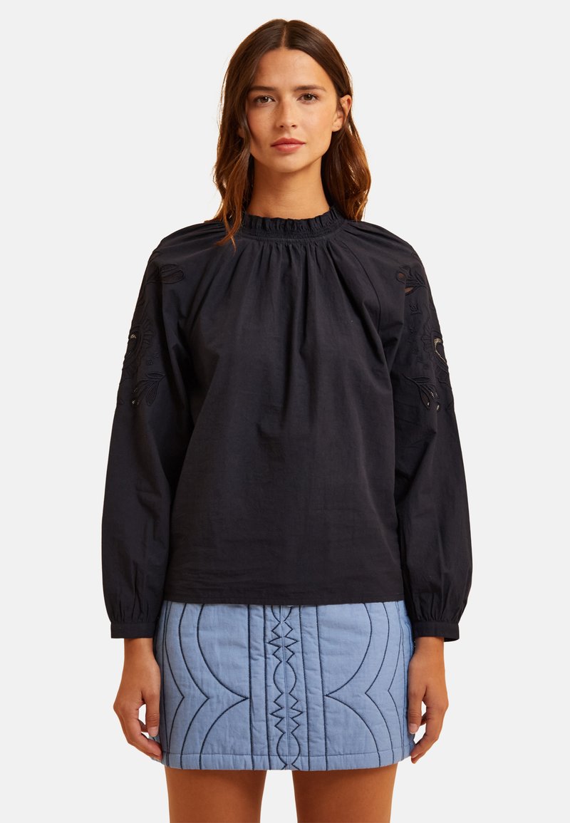 Blusa nera in cotone con scollatura arricciata e dettagli floreali ricamati sulle maniche, abbinata a una gonna trapuntata blu chiaro.