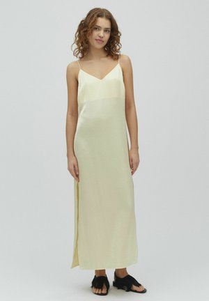 Kobieta o kręconych włosach, ubrana w jasnofioletową sukienkę maxi na cienkich ramiączkach i czarne sandały z piórkowymi ozdobami, stojąca na jednolitym tle.