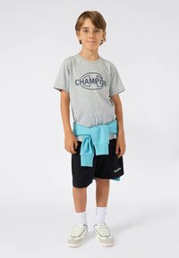 Berniukas stovi, vilkėdamas pilką Champion marškinėlį, juodas šortus, baltus sportinius batelius, baltas kojines ir mėlyną džemperį, surištą aplink liemenį.