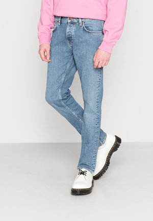 Persona que lleva vaqueros azules claros, zapatos blancos con cordones y suelas negras gruesas, y una camisa de manga larga rosa, de pie con las piernas cruzadas.