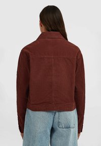Woman with long brown hair wearing a brown corduroy jacket and light blue denim jeans, shown from the back against a plain background.