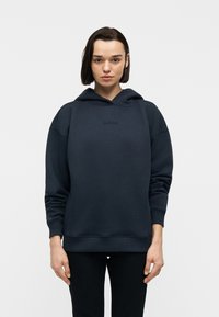 Νεαρό άτομο με κοντά σκούρα μαλλιά φορά μια σκουρόχρωμη μπλε ζακέτα με διακριτικό λογότυπο και μαύρο παντελόνι, στέκεται μπροστά σε μια απλή λευκή επιφάνεια.