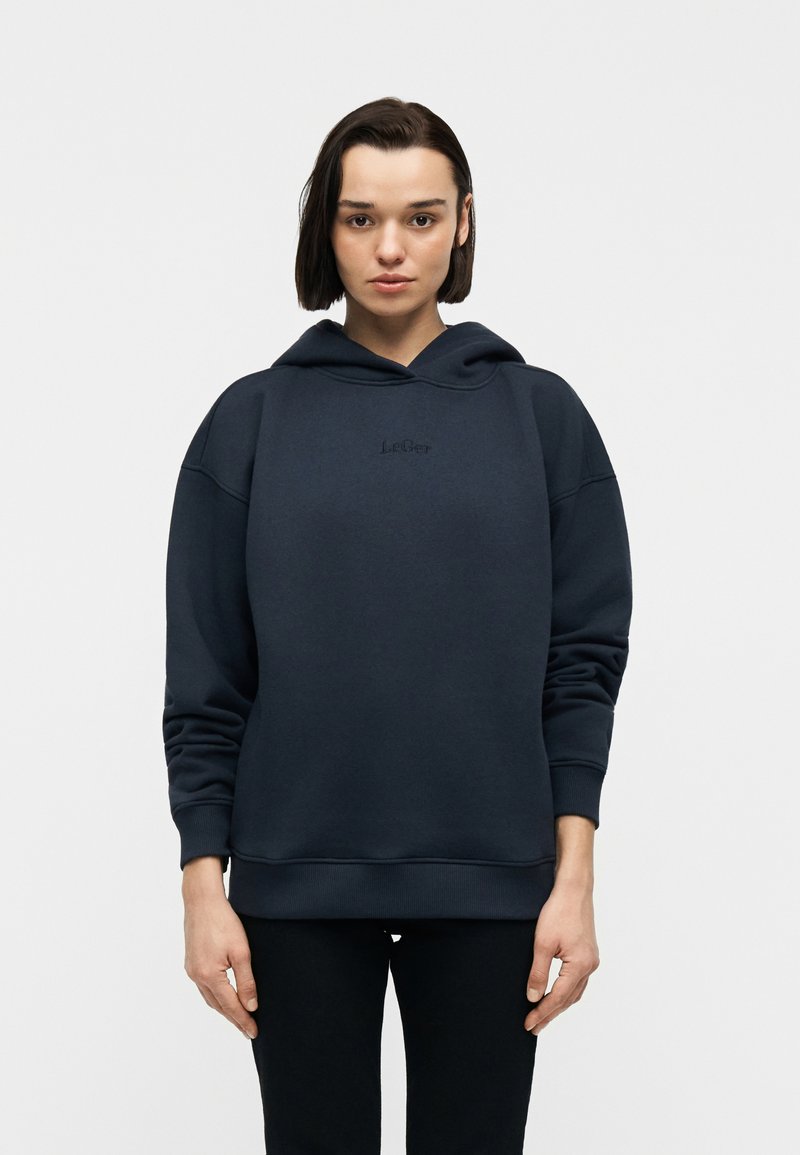 Νεαρό άτομο με κοντά σκούρα μαλλιά φορά μια σκουρόχρωμη μπλε ζακέτα με διακριτικό λογότυπο και μαύρο παντελόνι, στέκεται μπροστά σε μια απλή λευκή επιφάνεια.