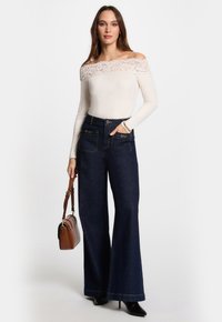 Weißes Oberteil mit schulterfreiem Schnitt und Rippstruktur, verziert mit Spitze, kombiniert mit hochwaistigen, dunklen, weit ausgestellten Jeans und einer kleinen braunen Tasche.