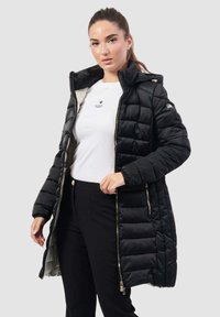 Giacca a vento nera con design trapuntato, cappuccio, chiusura con zip e tasche laterali. Indossata sopra una maglietta bianca e pantaloni neri, con dettagli dorati lucidi.