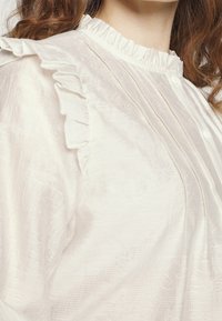 Blusa bianca con collare a volant, tessuto strutturato e dettagli con bottoni, caratterizzata da plissettature verticali e delicati motivi sulla superficie.