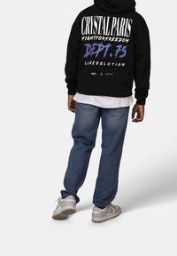 Musta huppari, jossa on graafinen teksti selässä, yhdistettynä vaaleansinisiin farkkuihin ja harmaisiin lenkkareihin. Hupparissa on rento istuvuus.