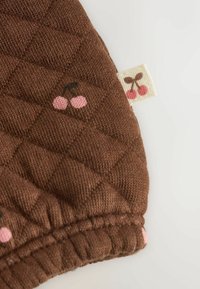 Pikowany brązowy materiał w kratkę, z różowymi motywami wiśni i małą metką z wiśniami. Miękka faktura, zaokrąglone krawędzie.