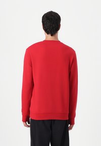 Sudadera roja de cuello redondo, de manga larga, con textura suave. Puños y dobladillo acanalados, detalle de costura mínima en la parte trasera. Pantalones negros.