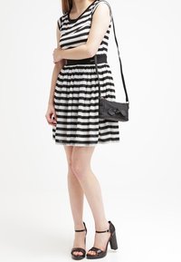 Robe sans manches à rayures noires et blanches avec une taille ajustée, jupe évasée, et un sac bandoulière noir. Sandales à talons hauts noires à bout ouvert.