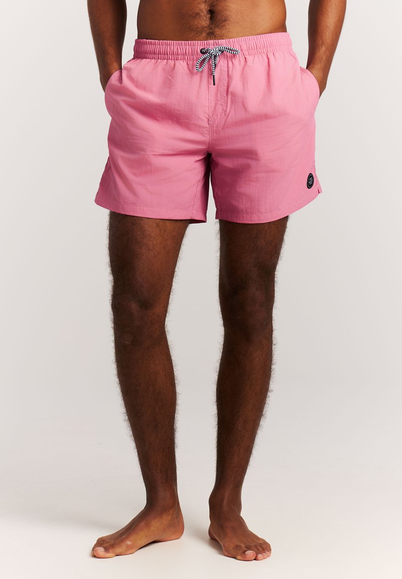 Man draagt roze zwemshort met zwart-wit koord, handen in de zakken, staat op blote voeten tegen een effen achtergrond.