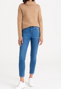 Jasnobrązowy, prążkowany sweter z wysokim kołnierzem, zestawiony z niebieskimi spodniami slim-fit i białymi sneakersami. Casualowy strój o prostych elementach designu.
