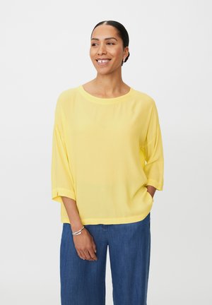 Żółta, lekka bluzka z okrągłym dekoltem i rękawami do łokcia. Połączona z niebieskimi szerokimi spodniami. Gładka tekstura, swobodny krój.
