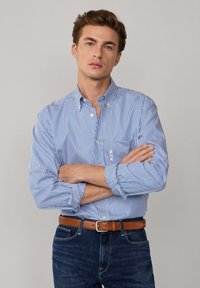 Joven con los brazos cruzados, vestido con una camisa de botones a rayas azul y blanca, cinturón marrón y jeans azul oscuro, contra un fondo liso.