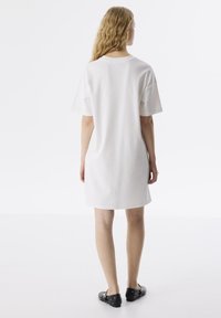 Witte katoenen t-shirt jurk met korte mouwen, ronde halslijn en een rechte snit. Model draagt zwarte platte schoenen. Minimalistisch ontwerp.