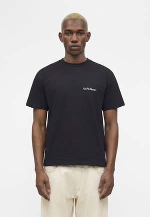 Rövidre festett szőke hajú férfi, fekete pólót visel, amelyen a mellkasán a „No Problemo” felirat látható, és világos bézs színű nadrágot, tiszta háttér előtt áll.