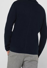 Navy textured sweater with a collar, long sleeves, and a straight hemline. Back seam detail visible, paired with grey trousers.