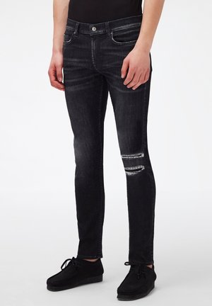 Czarne dżinsy slim fit z przetartymi rozdarciami na prawym kolanie, noszone z czarnymi butami i czarną bluzką.