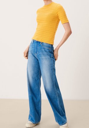 Person iført en tekstureret gul kortærmet top, vidtbenede blå jeans og hvide sneakers mod en ensfarvet neutral baggrund.