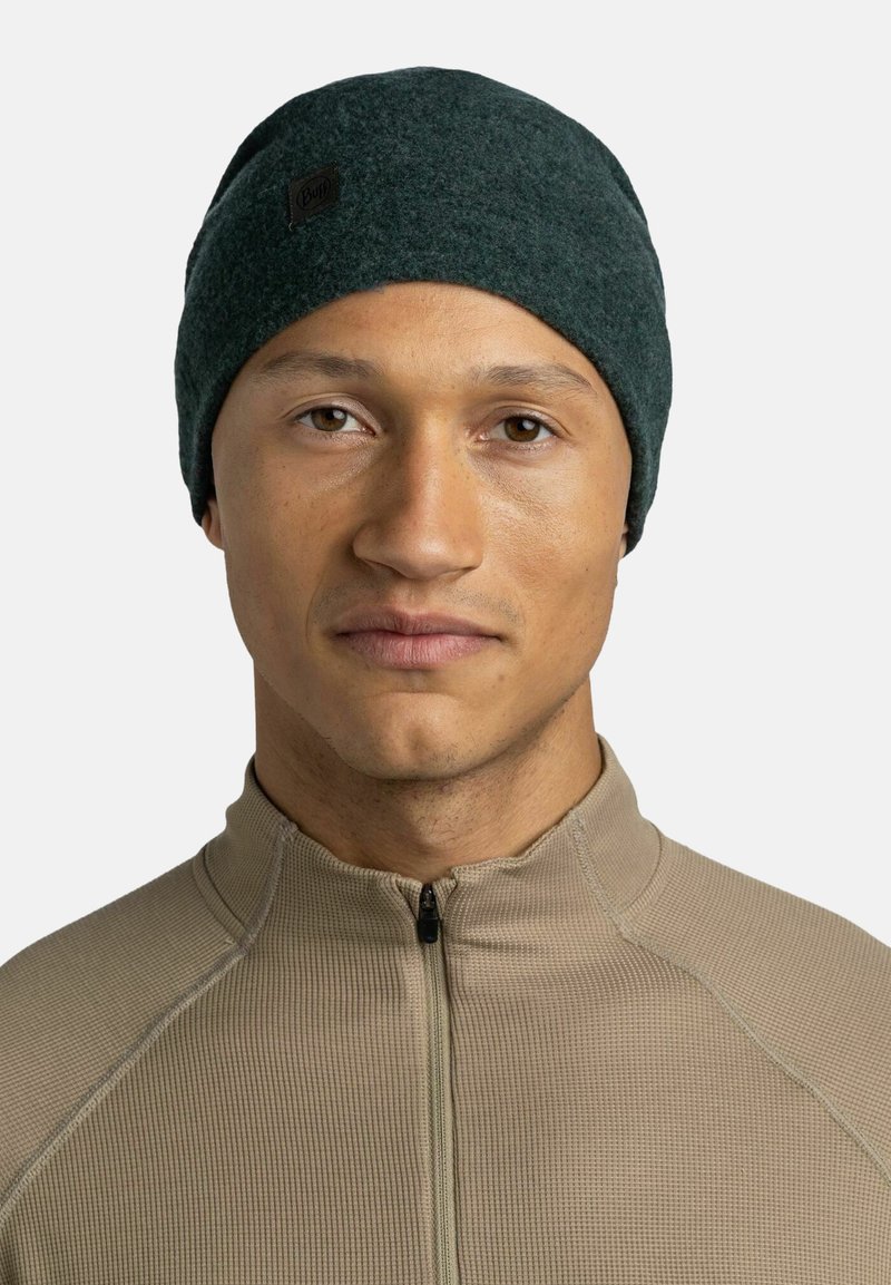 Dark green wool beanie with a folded cuff and a brown logo patch, worn by a person in a beige zip-neck top. Smooth texture.