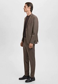 Man wearing brown suit with black shirt and black leather loafers, standing in profile on a plain light background.
