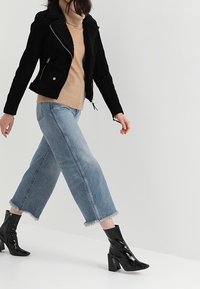 Giubbotto da motociclista in ecopelle scamosciata nera con colletto in shearling, dolcevita beige, jeans in denim a gamba larga e rovinati, e stivaletti neri in vernice alla caviglia.