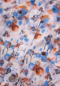 Bloemendessinfabric met tinten blauw, oranje en wit. Details omvatten kleine knopen en een gladde textuur.