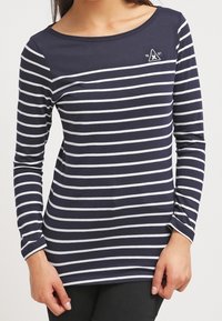 Woman wearing a navy blue long-sleeve shirt with white horizontal stripes and a small white emblem on the upper chest.