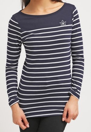 Vrouw in een marineblauwe lange mouwen shirt met witte horizontale strepen en een klein wit embleem op de bovenborst.