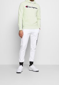 Světle zelená mikina s kapucí a logem "Champion", doplněná o bílé tepláky s čern ými akcenty. Obuv: bílé tenisky.