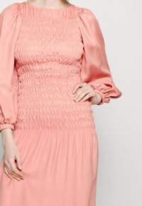 Vaaleanpunainen smokkihenkinen mekko pitkillä puffihihnoilla ja kuvioidulla vartalolla, jossa on liehuva hame ja sileä kangas.