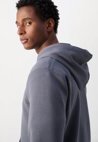 Siv hooded sweatshirt z glatko teksturo, opremljen s kapuco z vrvico in neizrazitimi šivi. Obleka je nošena in prikazuje stranski profil.