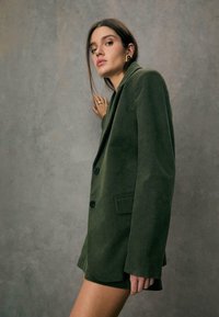 Blazer en velours côtelé vert foncé avec des revers crantés, deux boutons et poches latérales. La texture est côtelée, lui conférant une apparence distincte.
