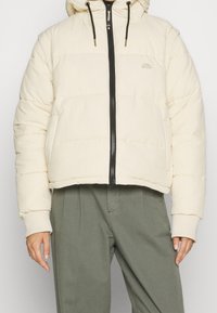 Chaqueta corta acolchada en beige claro, con cremallera negra y capucha con cordón. Tejido suave con patrón acolchado y bolsillos laterales.