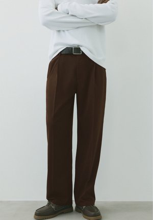 Personne portant un pantalon marron à jambes larges, une ceinture noire, des chaussures marron foncé et une chemise blanche à manches longues, les bras croisés.