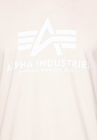 Béžové bavlněné tričko s výstřihem do V, na kterém je bíle vytištěné logo a text "ALPHA INDUSTRIES KNOXVILLE, TENNESSEE, U.S.A."