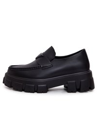 Mocassin en cuir noir avec semelle épaisse à crampons et détail de bride sur le dessus, présenté de profil sur fond blanc.