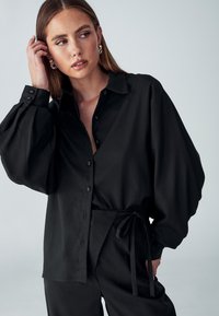 Camicia nera oversize con bottoni, maniche a palloncino e una texture liscia, abbinata a pantaloni a vita alta coordinati con una cintura da annodare.