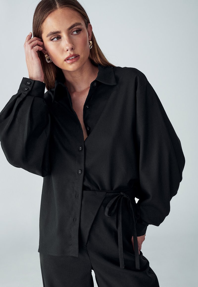 Camicia nera oversize con bottoni, maniche a palloncino e una texture liscia, abbinata a pantaloni a vita alta coordinati con una cintura da annodare.