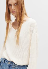 Pullover bianco in maglia strutturata con scollatura a V e spalle scese, abbinato a jeans di denim. I lunghi capelli lisci cadono sulle spalle.