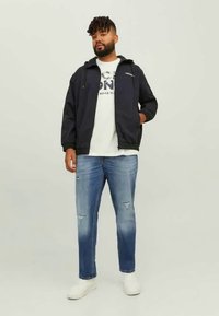 Ναυτική ζακέτα με κουκούλα, λευκό graphic t-shirt από κάτω, σκισμένα μπλε τζιν και λευκά αθλητικά παπούτσια, standing σε ουδέτερο φόντο.