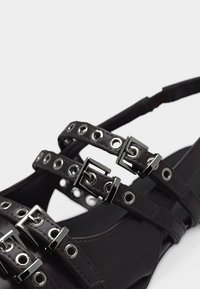 Μαύρο σανδάλι με λουράκια, τρία ρυθμιζόμενα λουράκια με αγκράφες, με ασημένιες τρύπες και τετράγωνες μεταλλικές αγκράφες σε απλό φόντο.