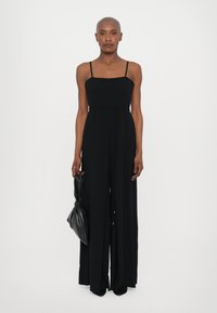 Black jumpsuit with a square neckline, thin straps, and a front slit. Paired with a textured black handbag and pointed black shoes.