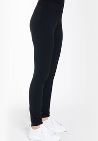 Zwarte leggings van een gladde, rekbare stof, met een hoge tailleband en een taps toelopende pasvorm, gecombineerd met witte sneakers op een neutrale achtergrond.