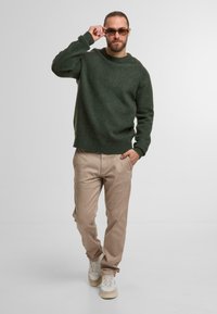 Groene wollen trui, beige broek en lichte sneakers. De trui heeft een ronde hals en geribbelde manchetten, wat zorgt voor een gestructureerd uiterlijk.
